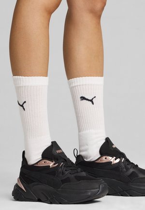 Ben iført hvide Puma mellemhøje sokker og sorte Puma sneakers med rosaguld accenter, stående på en grå baggrund.