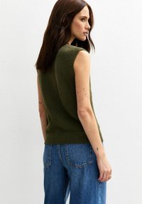 Pull en tricot sans manches vert olive avec une texture douce, un ourlet côtelé et une silhouette ajustée, associé à un jeans en denim bleu taille haute.