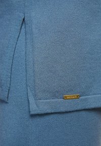 Un tissu tricoté bleu présente une surface texturée avec une fente sur le côté. Une petite étiquette dorée est cousue, indiquant la marque "Selmark".