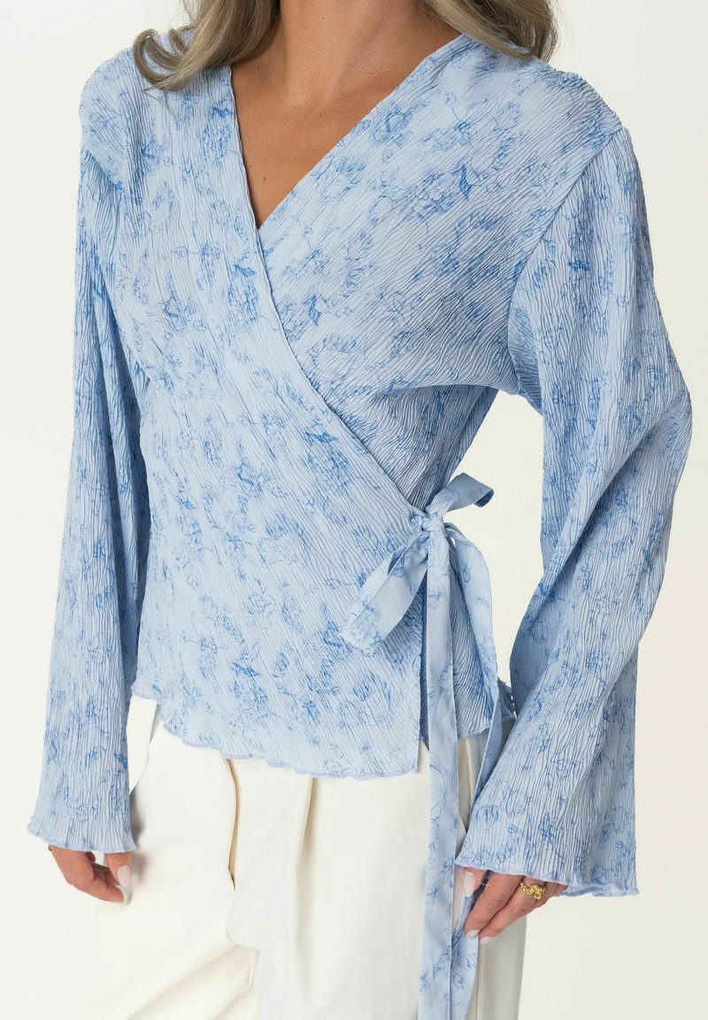 Helles blaues Wickeloberteil mit ausladenden Ärmeln, strukturierter Stoff mit floralem Muster und Bindegurt in der Taille. Sanftes, fließendes Design.