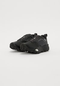Chaussures de sport noires avec un dessus texturé, des lacets proéminents, une semelle extérieure robuste, et des détails de logo sur le côté et le talon.