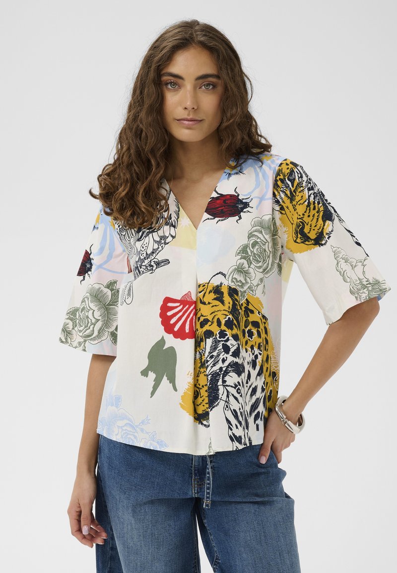 Ženska z kodrastimi lasmi nosi barvno bluzo z V-izrezom z živalskimi in cvetličnimi potiski ter modre kavbojke, stoji z eno roko v žepu.