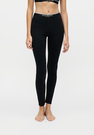 Žena, ktorá má na sebe čierne priliehavé legíny a zodpovedajúci čierny crop top s pásom s logom Emporio Armani, stojaca bosá na jednofarebnom pozadí.