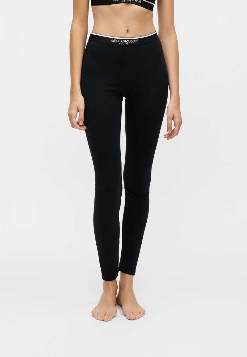 Femme portant des leggings noirs moulants et un crop top noir assorti avec une ceinture à logo Emporio Armani, debout pieds nus sur un fond uni.