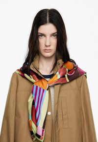 Bufanda de seda con patrones vibrantes en rojo, naranja, verde, azul y púrpura, anudada al cuello de una chaqueta marrón.