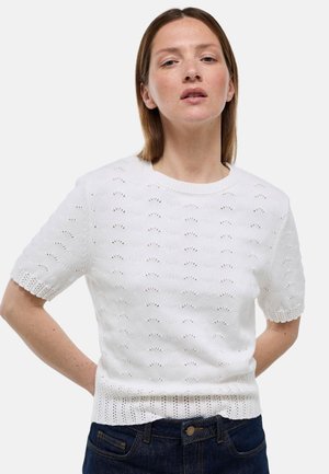 Femme à la peau claire et aux cheveux raides portant un haut en maille blanc à manches courtes avec un motif ajouré et un jean bleu foncé, debout les mains dans le dos.