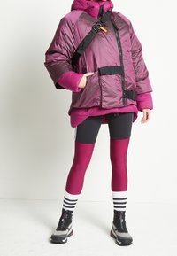 Puffermantel i skiftande lila nyanser, med huvtröja och frontdragkedja. Kombinerad med svarta shorts och magenta leggings som har vita randiga strumpor och stövlar.