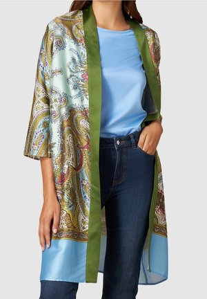 Kobieta ubrana w jasnoniebieską koszulę, ciemnoniebieskie dżinsy oraz wzorzysty jedwabny kimono z zielonym obszyciem, stojąca z jedną ręką w kieszeni.