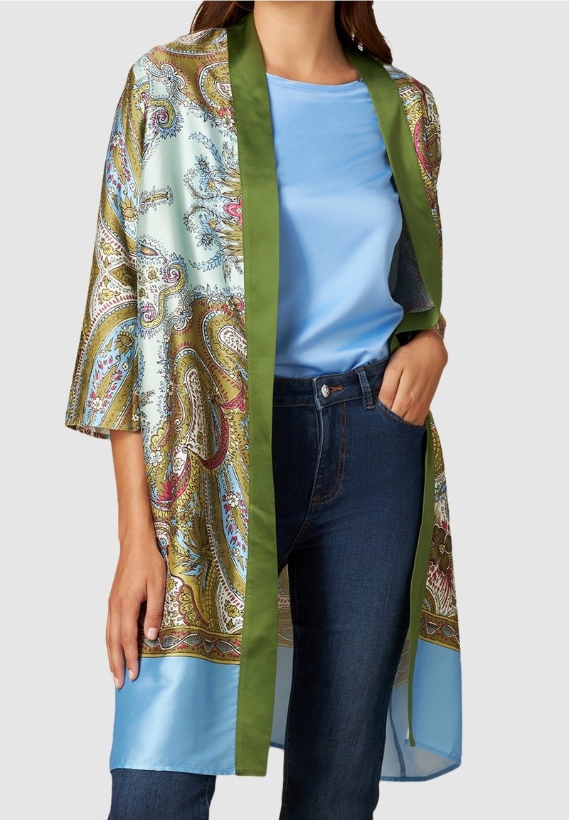 Femme portant une chemise bleu clair, un jean bleu foncé et un kimono en soie à motifs avec une bordure verte, debout avec une main dans la poche.