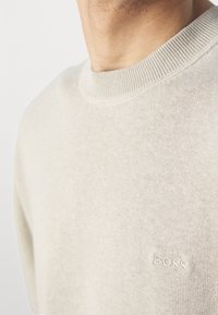 Pull léger beige avec un col rond, des poignets côtelés et un logo "BOSS" brodé sur la poitrine. Texture lisse, tissu tricoté.