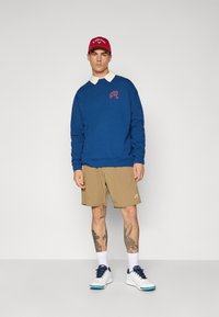 Sweatshirt azul com gola branca e logótipo bordado; calções caqui; ténis brancos com detalhes azuis; boné vermelho. Tatuagens visíveis nas pernas.