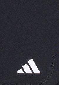 Logo Adidas blanc avec trois bandes diagonales sur un tissu noir près d'une couture cousue.