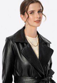 Cappotto trench in pelle nera con ampio colletto, vita cintata e dettagli sulle spalle. Il modello indossa una maglietta chiara e una serie di collane d'oro sovrapposte.
