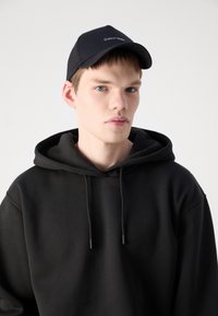 Hanorac negru cu o glugă mare și șnururi de strângere, purtat sub o șapcă neagră cu broderie albă a logo-ului "Calvin Klein".