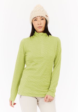 Žena u svijetlozelenom dugom puloveru teksture s pola zatvarača i kremastom pletenom kapom stoji na bijeloj pozadini.