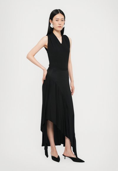 Black sleeveless dress with a V-neckline and asymmetric hem. Smooth fabric with a flowy texture. Model wears pointed black mules.