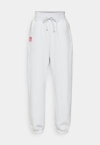 Hvit joggebukse med høy midje, elastiske mansjetter og et rødt logo-lapp på venstre side. Laget av et mykt, teksturert stoff.