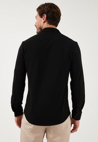 Camisa negra de manga larga con una textura sutil, con puños de botón y un dobladillo curvo, vista desde la parte trasera sobre un fondo neutro.