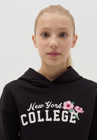 Sudadera negra con la inscripción "New York COLLEGE" en color blanco, adornada con flores bordadas rosas en el lado derecho. Diseño sencillo, capucha con cordón.
