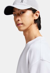 Casquette blanche avec un design perforé et un bord sombre, portée avec un sweat-shirt à col rond blanc. Accent sur le matériau et la silhouette épurée.