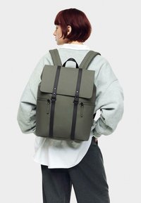 Sac à dos vert avec une forme structurée, matériau semblable au cuir, rabat supérieur, bretelles noires, deux zips à l'avant et détail de marque à l'avant.