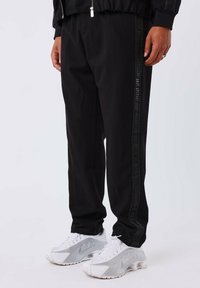 Zwarte joggingsbroek met een gestructureerde streep langs de zijkant, gemaakt van zachte stof, gecombineerd met witte sportschoenen.