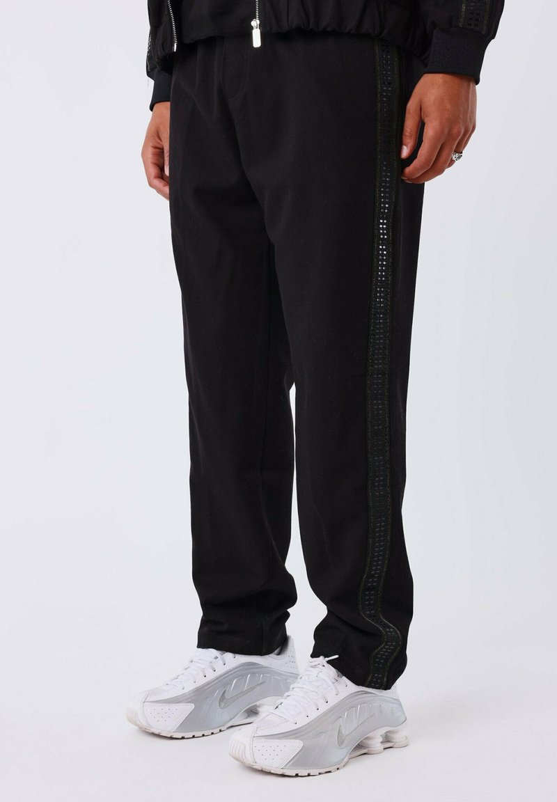 Zwarte joggingsbroek met een gestructureerde streep langs de zijkant, gemaakt van zachte stof, gecombineerd met witte sportschoenen.
