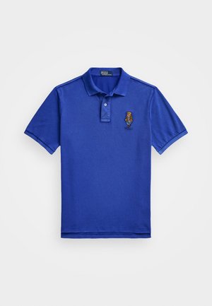 Camisa polo azul feita de algodão. Apresenta uma pala com dois botões, mangas curtas e um logótipo bordado no lado esquerdo do peito. Textura suave.