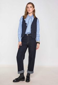 Gilet a righe navy sopra una camicia a righe blu chiaro con un papillon, abbinato a pantaloni in denim scuro e scarpe nere.