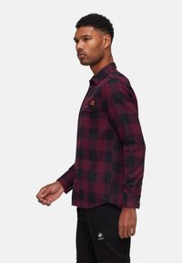 Uomo che indossa una camicia a quadri rossa e nera a maniche lunghe con bottoni e pantaloni neri, in piedi di profilo su sfondo bianco.