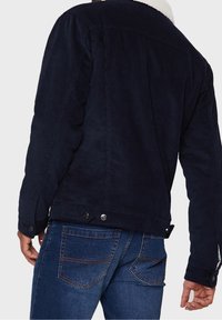 Giacca in velluto a coste blu marina con colletto in pile bianco, polsini con bottone e spalla a yoke. Indossata con jeans in denim scuro caratterizzati da cuciture a contrasto.