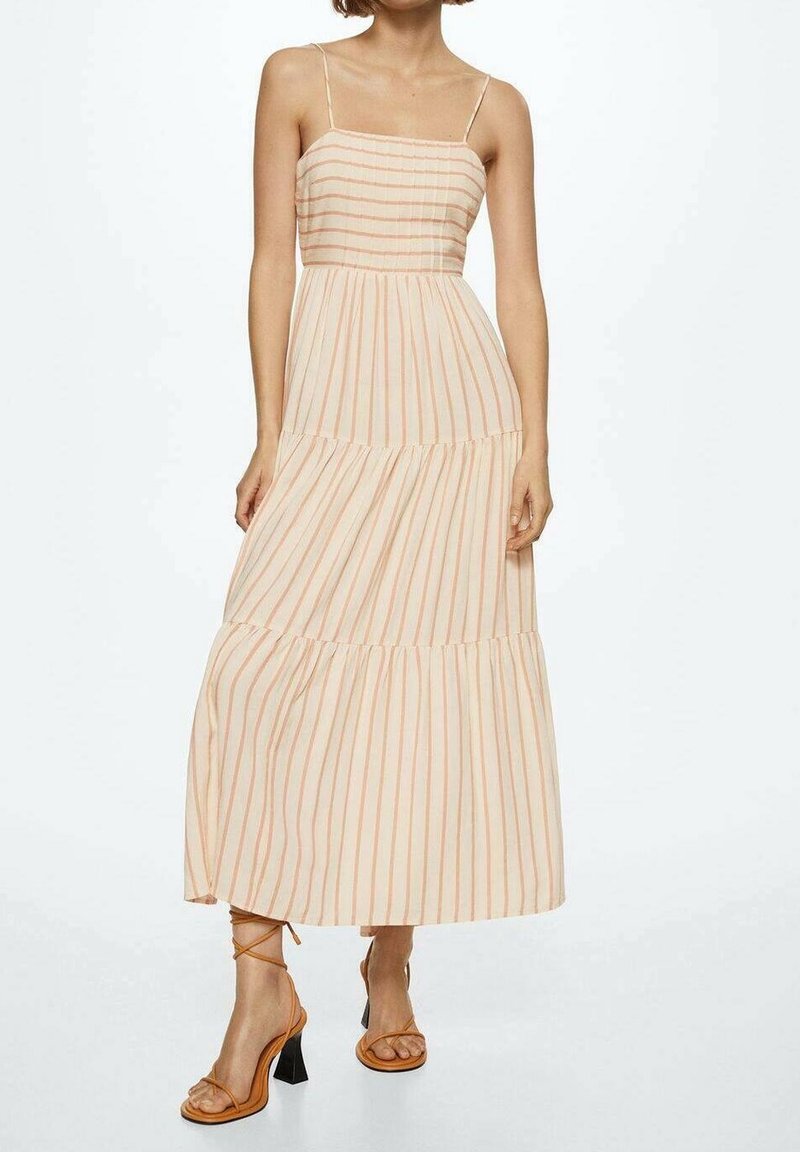Femme portant une robe longue sans manches beige clair avec de fines rayures verticales orange et des sandales marron à talons hauts et à lanières.