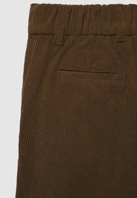 Pantalon marron en velours côtelé avec une taille élastique, passants de ceinture et une poche arrière passepoilée sur fond clair.
