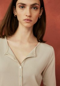 Beige ribbet top med en knaplukning ved halsen og lange ærmer. Stoffet er blødt med en tekstureret overflade.