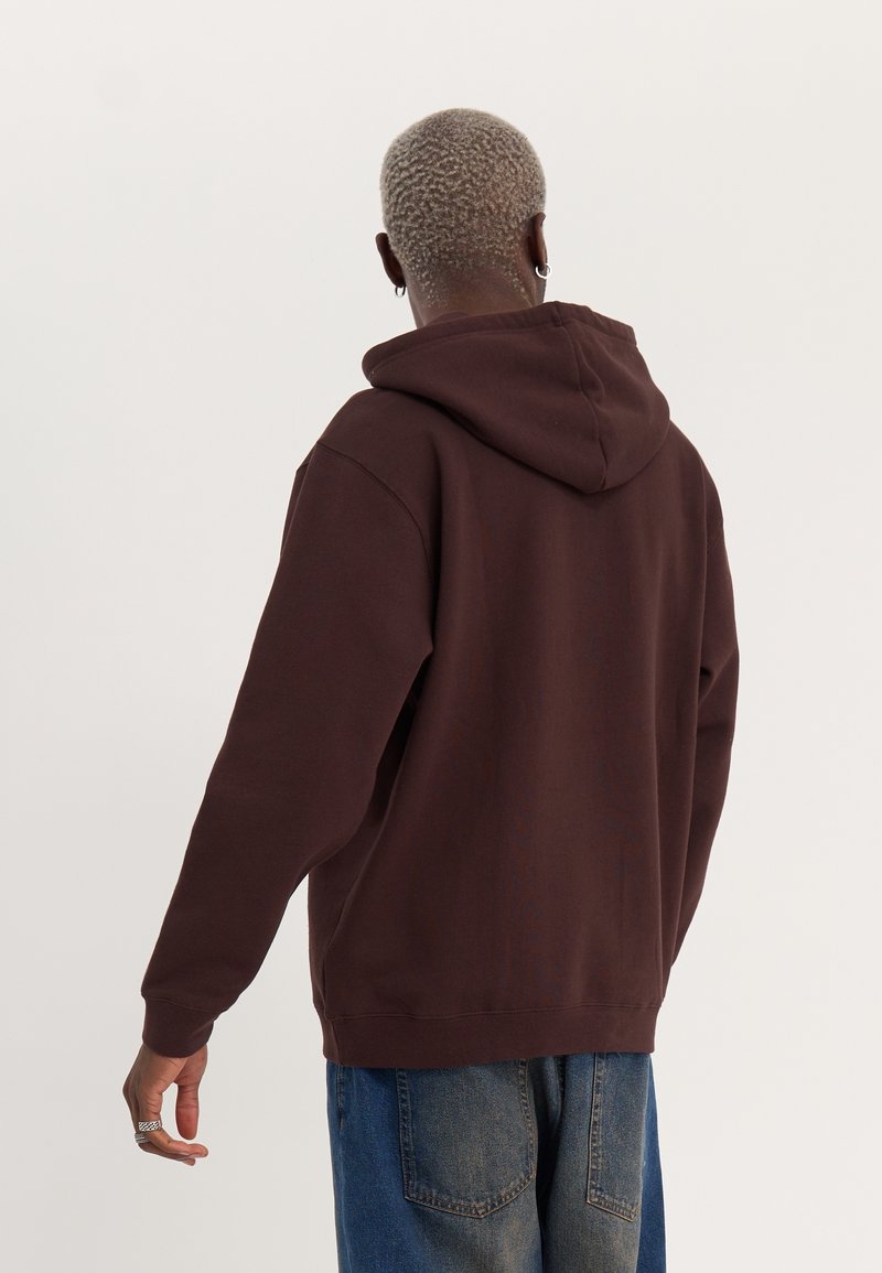 Personne aux cheveux blonds courts et bouclés, portant un sweat à capuche marron foncé et un jean bleu, vue de dos sur un fond uni.