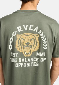Olijfgroene T-shirt met korte mouwen, voorzien van een grote gele tijgerafbeelding en witte tekst op de achterkant. De opdruk heeft een gebarsten textuur.