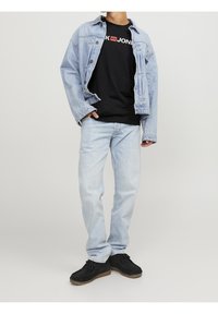 Denim jacka med ljus tvätt och stora fickor över en svart t-shirt med vit och röd logotyp. Ljusblå jeans och svarta skor.