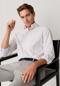 Camicia bianca button-up con motivo a quadri chiari, maniche lunghe e colletto classico. Indossata con pantaloni grigi e una cintura scura, seduto su una sedia nera.