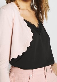 Ljusrosa cardigan med scallopade kanter över en svart spetscami, kombinerat med rosa byxor, vilket visar mjuka texturer och kontrasterande färger.