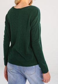 Zielony sweter z dzianiny o szerokim dekolcie, ściągaczu na dole i obniżonych ramionach, zestawiony z jasnoniebieskimi dżinsami. Miękka faktura i luźny krój.