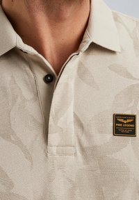 Polo beige en tissu texturé, avec un col détaillé et un bouton noir. Un logo emblématique PME Legend est brodé sur la poitrine.