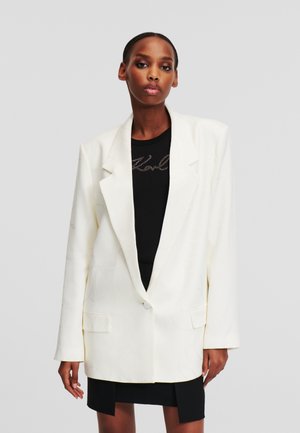 White blazer with a subtle textured pattern, single button closure, and two front pockets, layered over a black top, worn with a mini skirt.