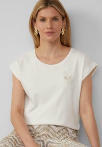 T-shirt blanc à manches courtes avec un design de ruche dorée sur la poitrine. Tissu doux avec un col rond. Le modèle porte un bas texturé et à motifs.