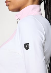 Balta ilgomis rankovėmis palaidinė su rožiniu apykakle ir sidabriniu užtrauktuku. Ant rankovės yra juodas logotipo užtasas. Lygus, elastingas audinys.