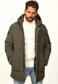 Olivgrüne Daunenjacke mit Kapuze, Reißverschluss und gerippten Bündchen; getragen über einem weißen Kabelstrickpullover und kombiniert mit hellblauen Jeans.