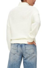 Biały sweter z dzianiny z wysokim kołnierzem, o luźnym kroju i prążkowanym wykończeniu. W połączeniu z jasnoniebieskimi dżinsami z tylnymi kieszeniami.