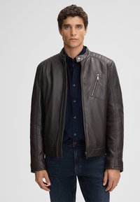 Braune Lederjacke mit Reißverschluss, gerippten Akzenten an den Schultern und einer Reißverschlusstasche auf der Brust. Über einem dunkelblauen Hemd getragen.