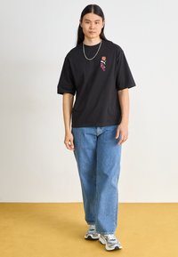 Svart bomullströja med korta ärmar och grafik fram, kombinerad med ljusblå denimjeans och grå atletiska skor.