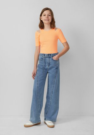 Neon oranje top met korte mouwen en uitgesneden schouders, gecombineerd met hoogwaardige, wijd-uitlopende lichtblauwe jeans en witte sneakers met beige accenten.
