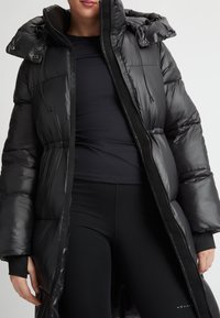 Zwarte pufferjas met glanzende afwerking, getailleerde taille, grote capuchon en ritssluiting. Gedragen over een donkere top en strakke zwarte leggings.